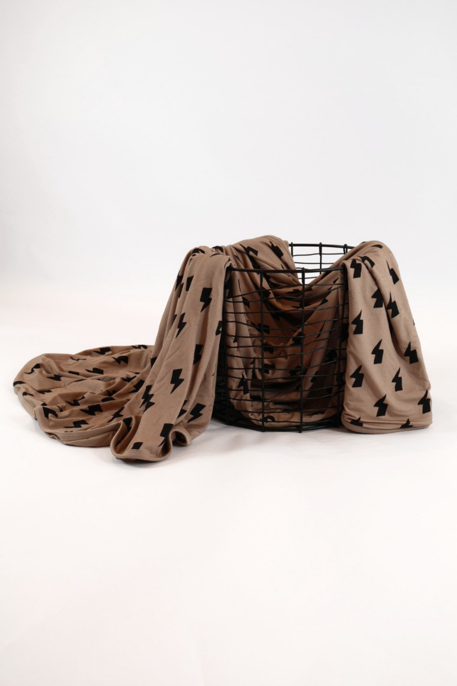 Black lightning bolts scattered across a warm taupe blanket with a warm taupe back accent and trim draped over a black wire basket on a white background.