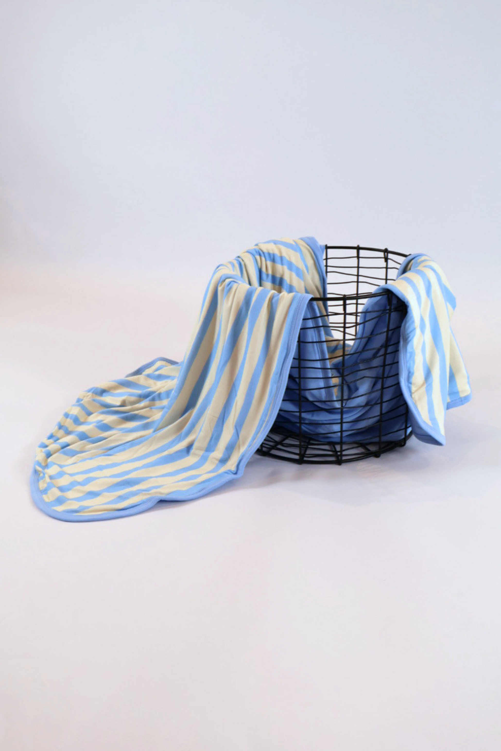 Our Coastal Stripes triple layer blanket features soft blue and cream vertical stripes with a complementary soft blue back accent and trim. It is shown featured over a black wire basket.