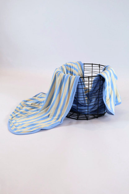Our Coastal Stripes triple layer blanket features soft blue and cream vertical stripes with a complementary soft blue back accent and trim. It is shown featured over a black wire basket.