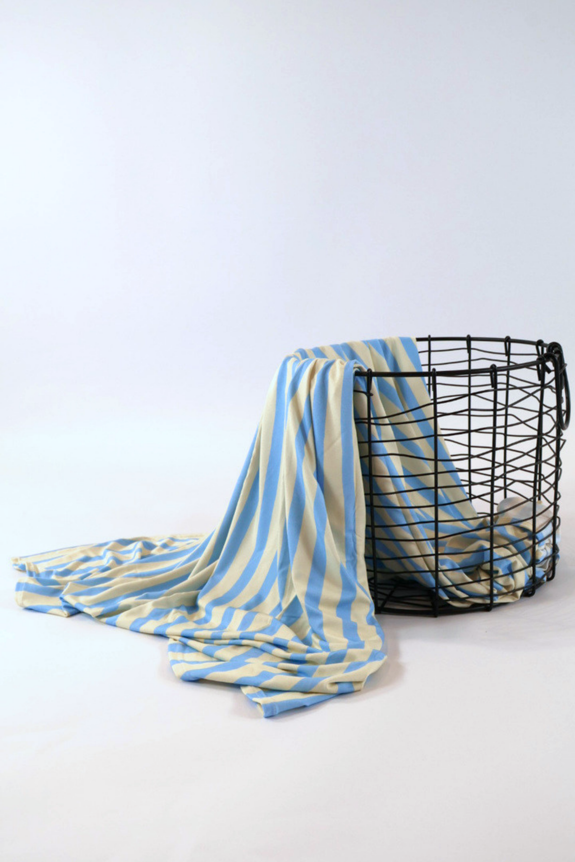 Our Coastal Stripes swaddle features soft blue and cream vertical stripes. It is shown featured over a black wire basket.