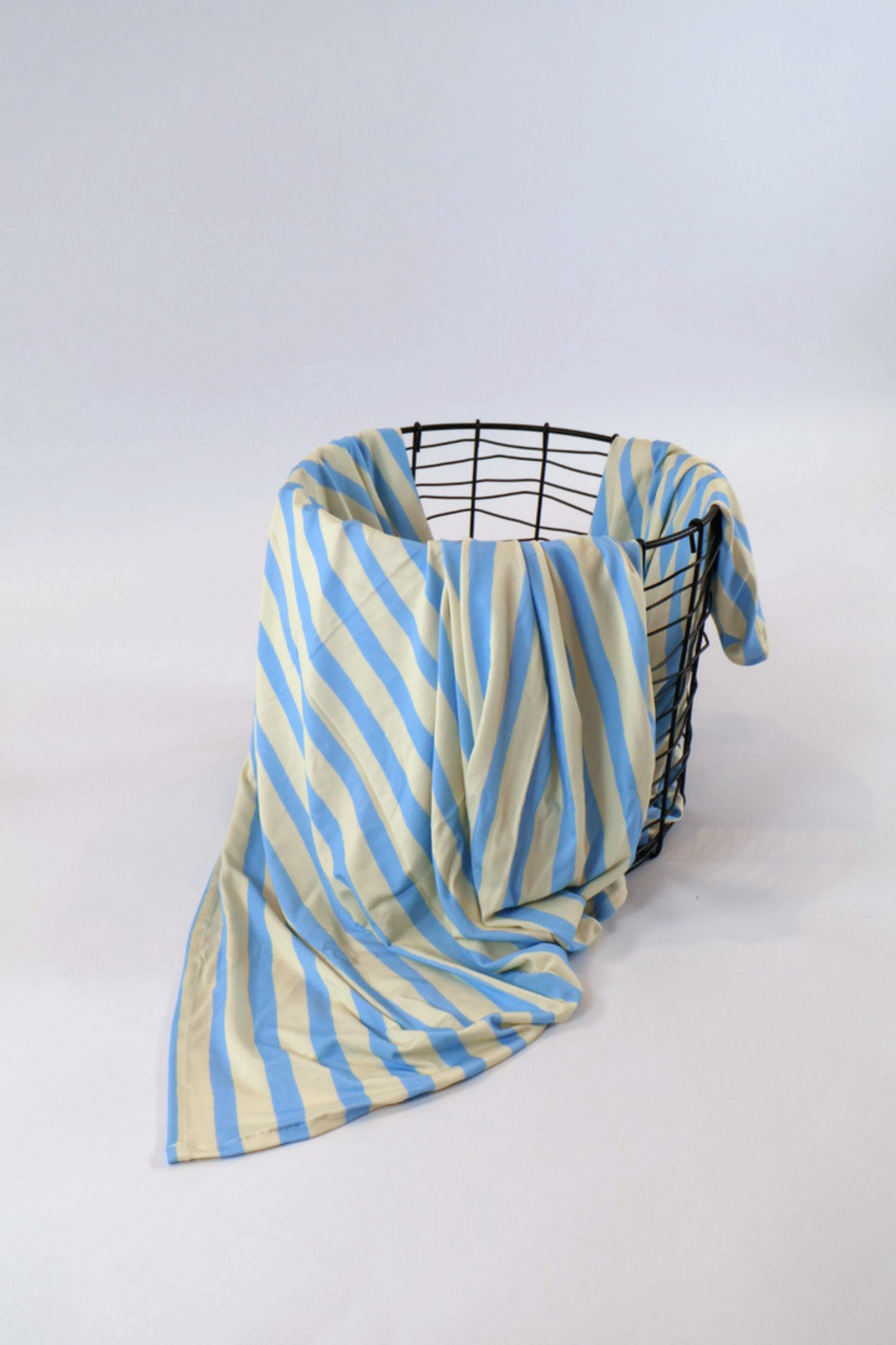 Our Coastal Stripes swaddle features soft blue and cream vertical stripes. It is shown featured over a black wire basket.