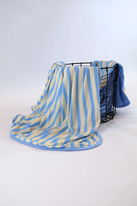 Our Coastal Stripes triple layer blanket features soft blue and cream vertical stripes with a complementary soft blue back accent and trim. It is shown featured over a black wire basket.